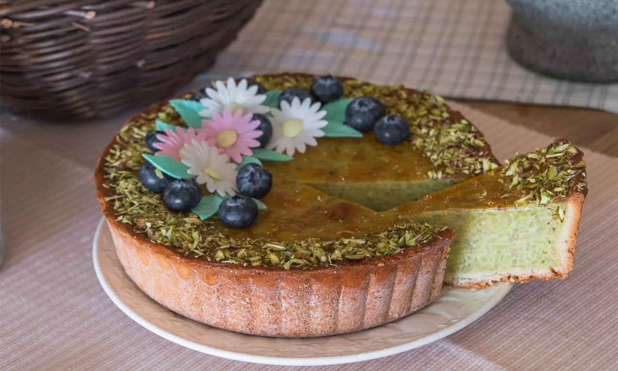 Limburský rýžový koláč se zlatohnědou křupavou okrajem, zelenkavou náplní, zdobený pistáciemi, borůvkami a cukrovými květinami na světlém talíři