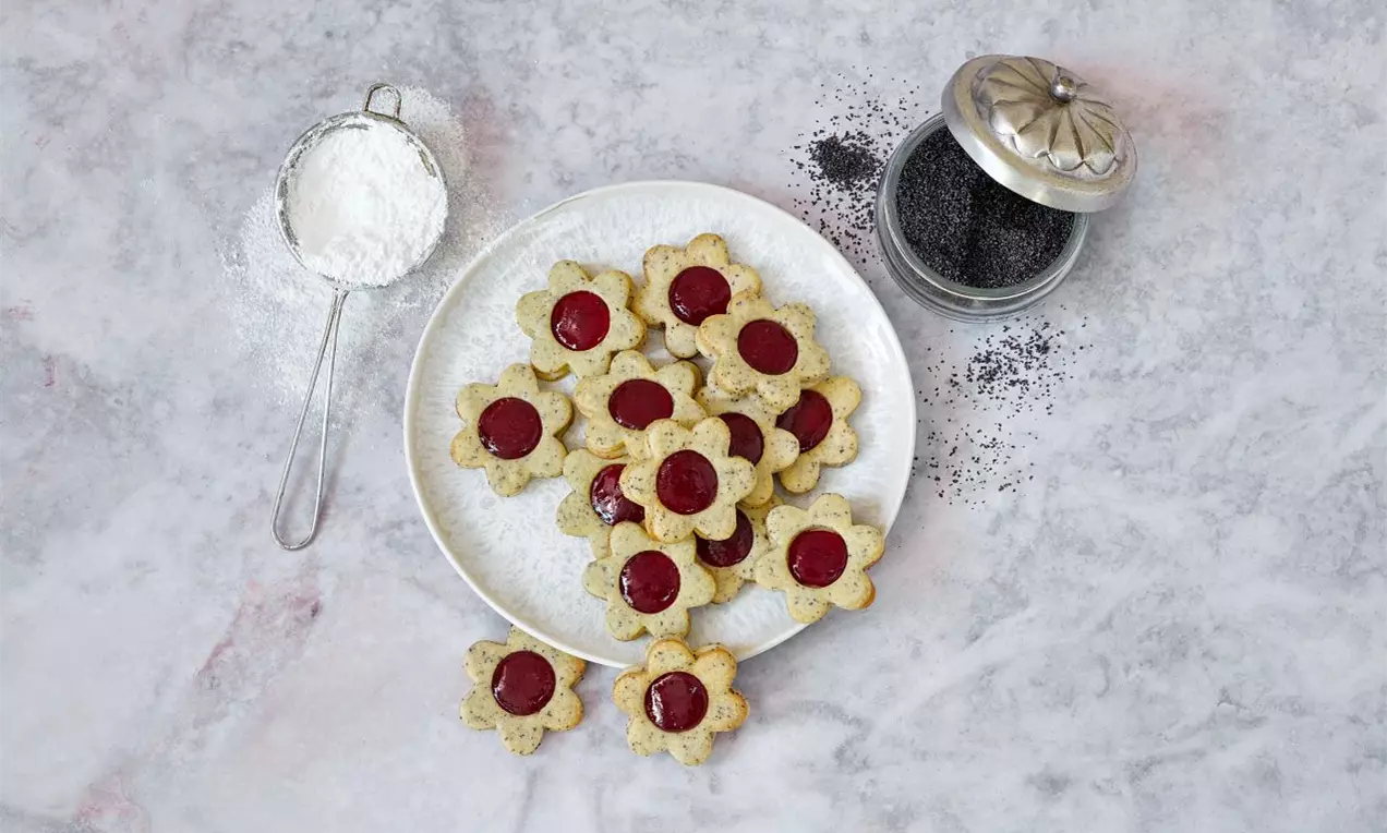 Makové linecké kytičky světle zlaté barvy s červeným středem uspořádané na bílém talíři, vedle sítka s moučkovým cukrem a sklenice máku