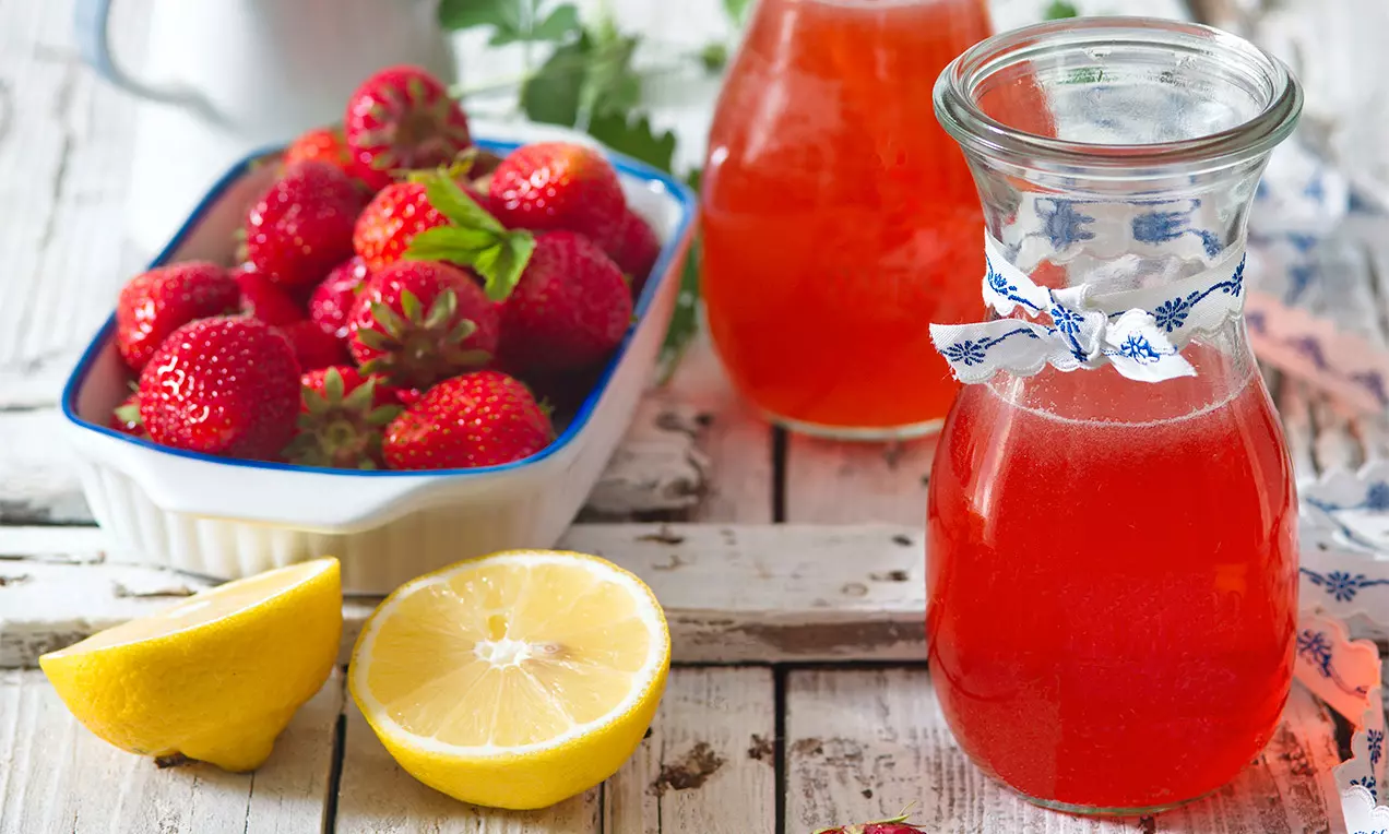 Jasně červený sirup ve skleněné karafě zdobené mašlí, vedle čerstvé jahody v bílé misce a půlky citronu na světlém dřevěném povrchu