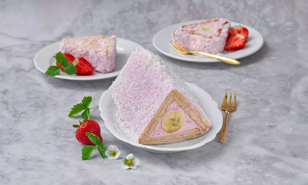 Trojúhelníkový řez s růžovou jahodovou náplní, ukrytou v kokosové posypce, uprostřed banán, podávaný na bílém talíři s čerstvými jahodami