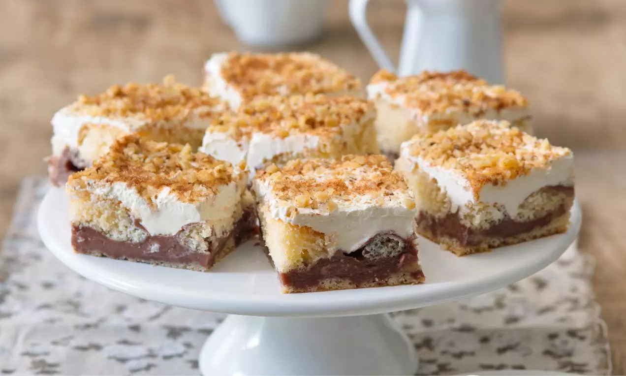 Čtvercové pudingové moučníky s vrstvou čokolády, světlého krému a zlatavých drobenkových posypů na bílé keramické míse na dekorativní látce