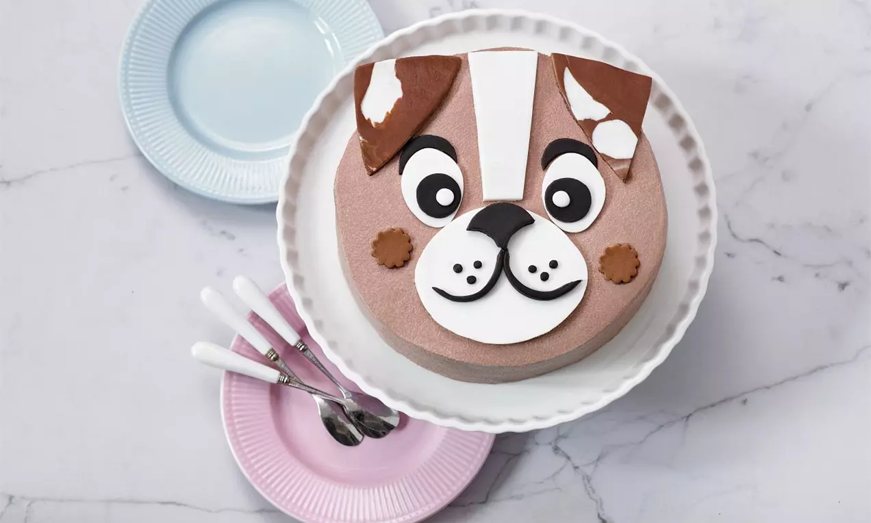 Tvarohový dort kulatého tvaru s obličejem psa z hnědé, bílé a černé polevy na bílém talíři, vedle barevných talířků a lžiček