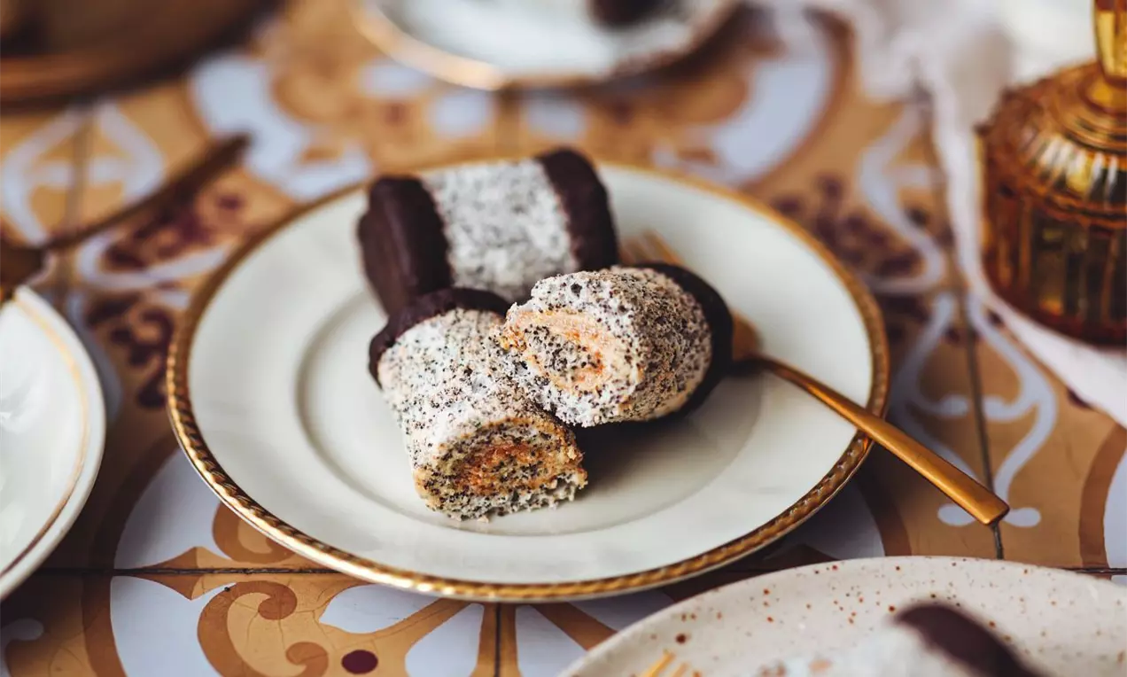 Makový rolovaný moučník plněný hnědým karamelovým krémem, politý tmavou čokoládou na koncích na talířku se zlatým okrajem a zlatou vidličkou