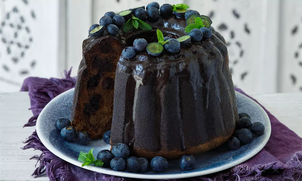 Kakaová bábovka plněná borůvkami, zdobená čerstvými borůvkami a borůvkovými lístky na bílém podkladu a fialovém šátku