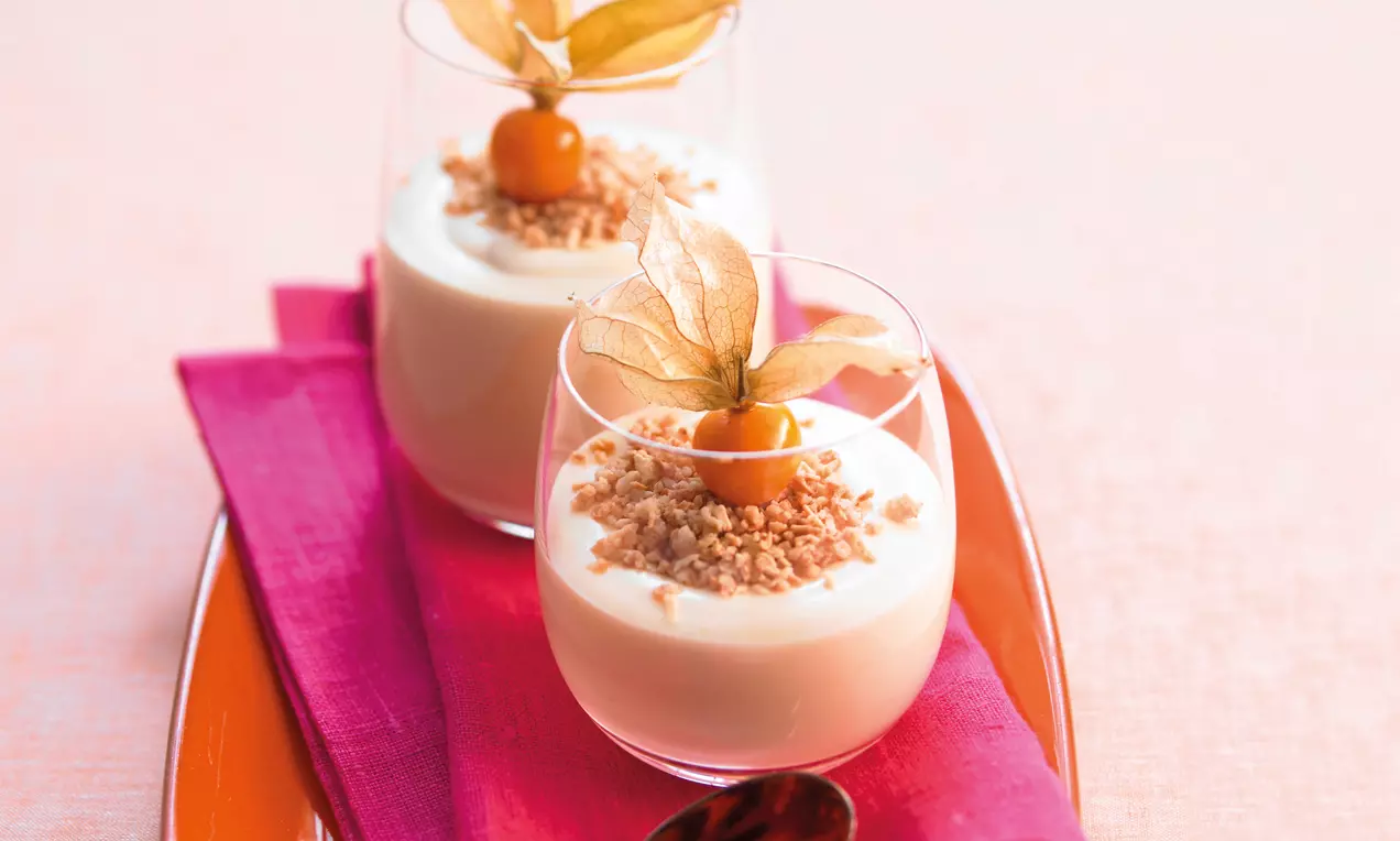 Krém světle smetanové barvy v průhledných skleničkách, posypaný drobnými oříškovými kousky a ozdobený physalis na růžovém ubrousku