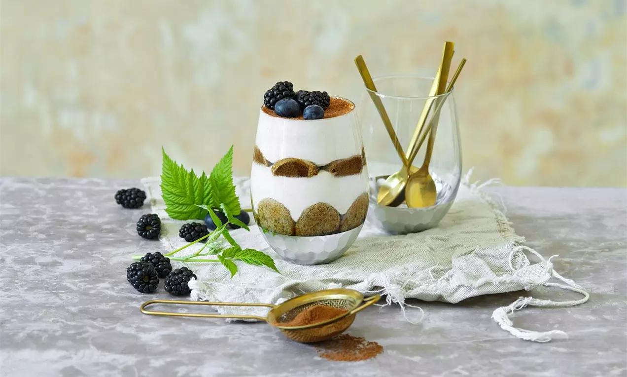 Sklenice s vrstveným dezertem z krémového bílého mascarpone, kakaového prášku, piškotů, ostružin a borůvek na šedém povrchu
