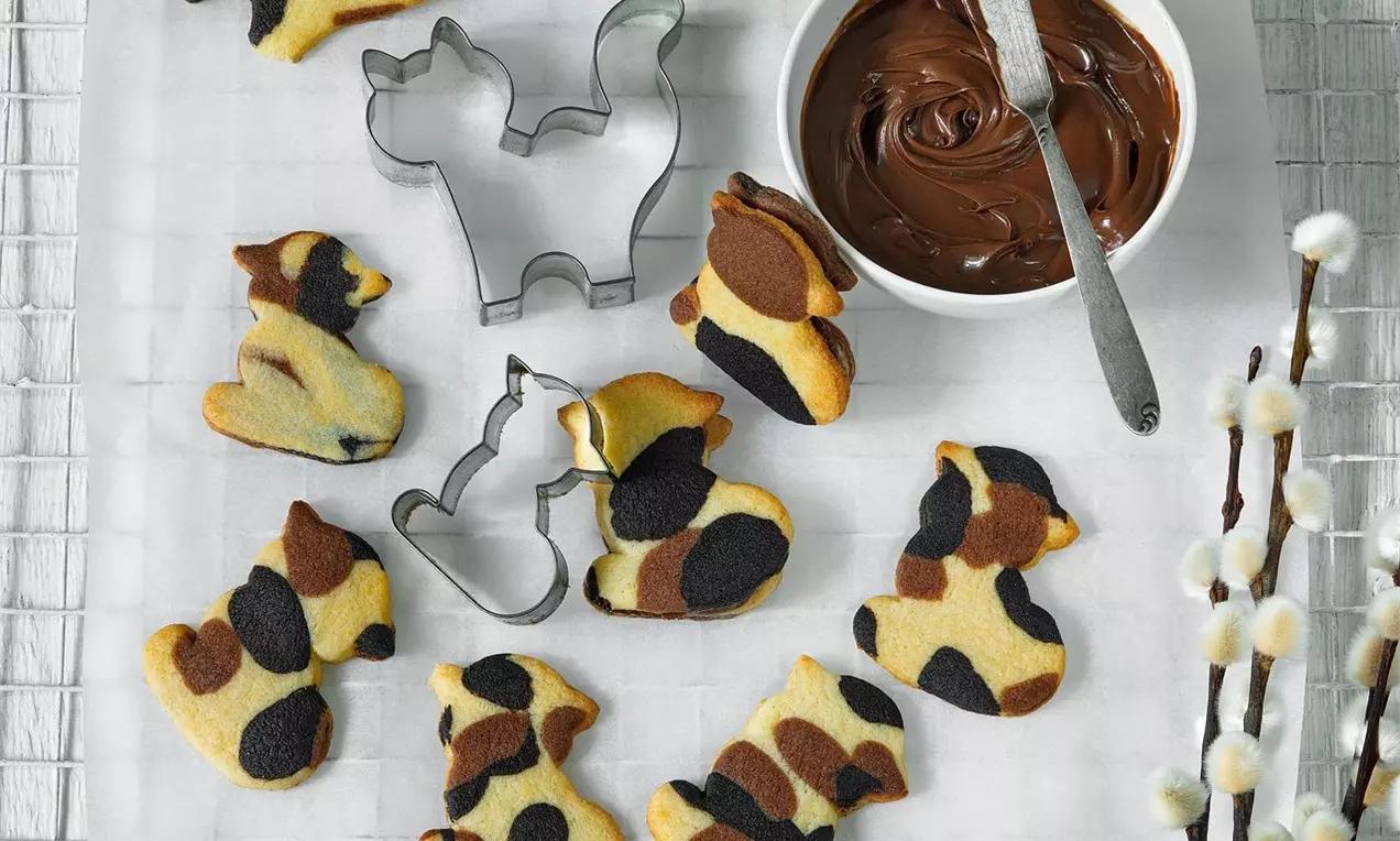 Sušenky ve tvaru sedících koček, máslové barvy s hnědými a černými skvrnami, naaranžované na bílém podkladu vedle vykrajovátek a misky s čokoládou