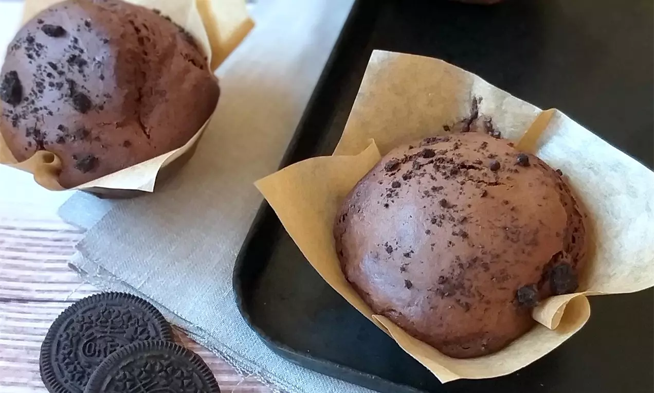 Čokoládové muffiny s tmavým, lehce popraskaným povrchem v hnědých papírových košíčcích, sypané drobky Oreo sušenek na černém podnosu