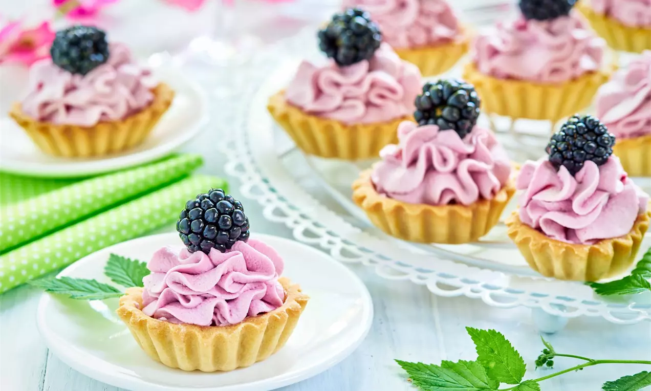 Mini tartaletky s borůvkovým krémem zdobené čerstvou ostružinou na bílem servírovacím a dezertním talířku