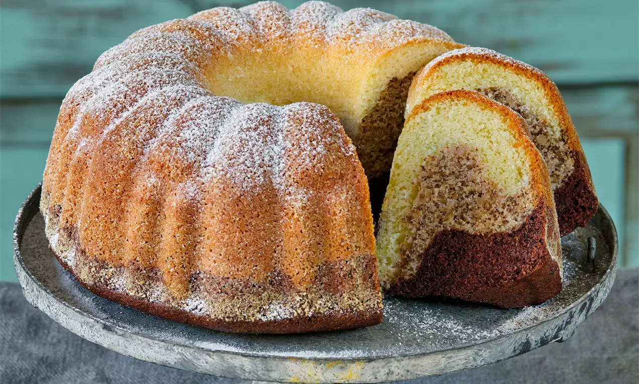 Farská bábovka ve tvaru rozevřeného věnce, zlatohnědá s mramorovanou strukturou, lehce posypaná cukrem, na rustikálním kovovém podnosu
