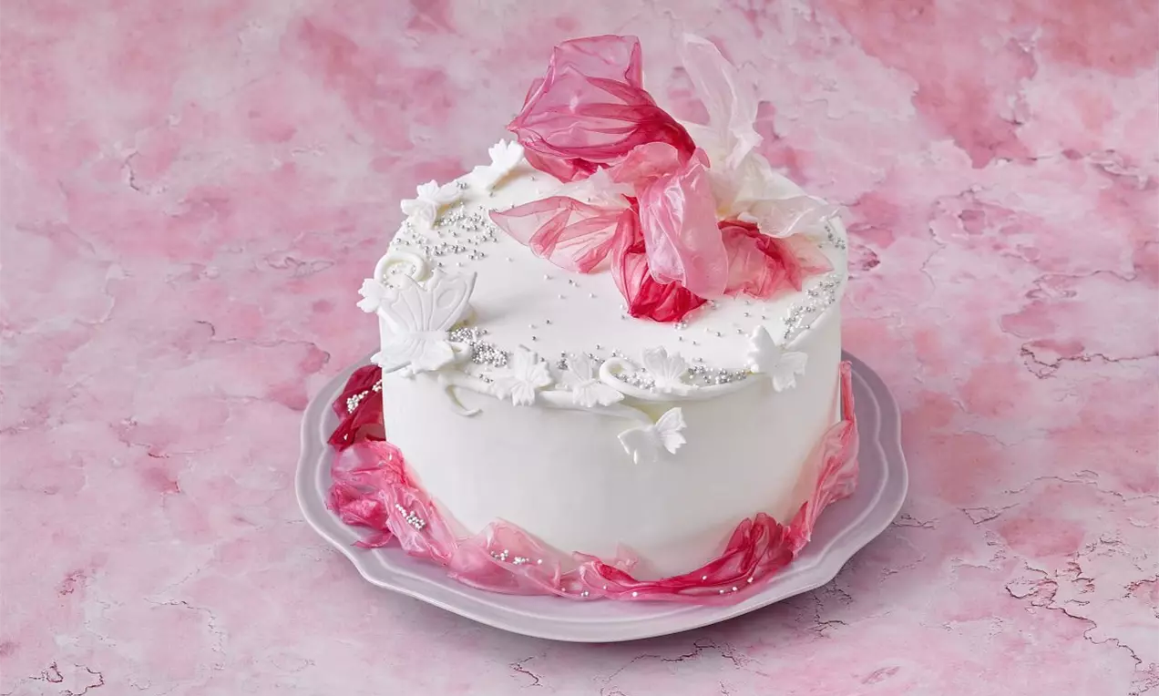 Bílý dort s hladkým povrchem, ozdobený růžovými a bílými květinovými dekoracemi na světlém talíři, položený na růžovém mramorovém pozadí