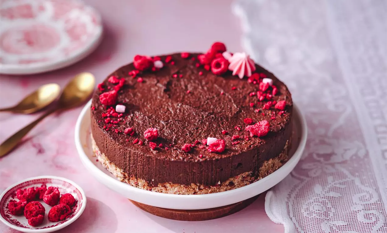 Čokoládový nepečený dort na světlém korpusu zdobený mrazem sušenými malinami na růžové pracovní desce
