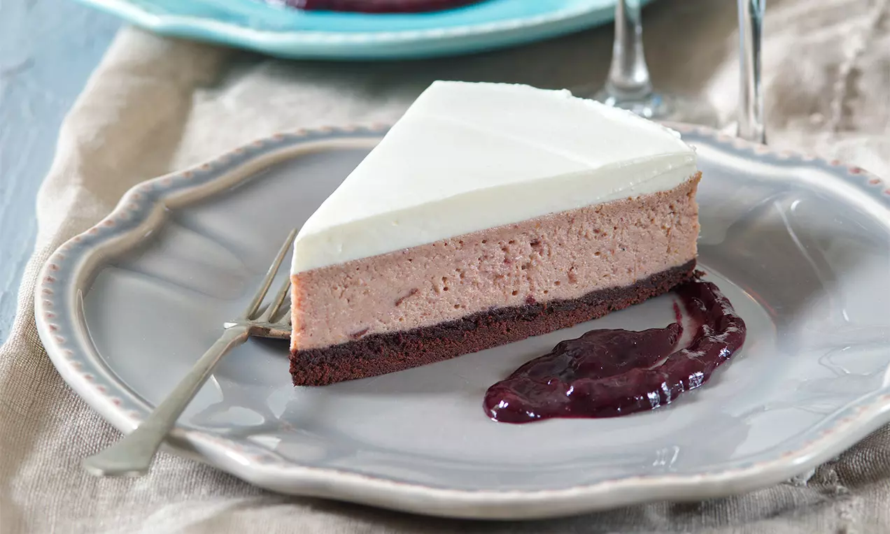 Krémový cheesecake s bílou vrstvou na čokoládovém základu, podávaný na vroubkovaném talíři s povidlovou omáčkou vedle kousku dortu