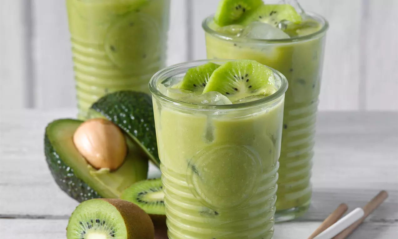 Zelený nápoj v žebrovaných sklenicích s plátky kiwi a ledem, vedle čerstvé avokádo s peckou a kiwi na světlém dřevěném povrchu
