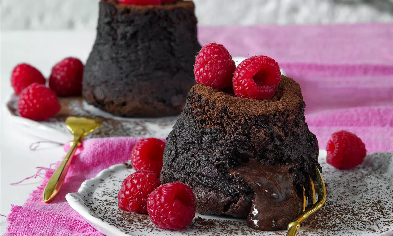 tmavě hnědé čokoládové suflé s tekutým středem na bílém talířku se zlatou vidličkou nazdobené čerstvými malinami