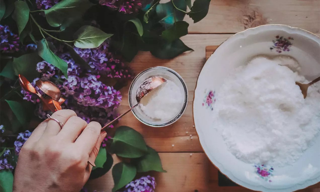 Bílý jemný prášek v porcelánové misce s květinovým motivem, vedle sklenička a bronzová lžička, okolí zdobí fialové květy šeříku