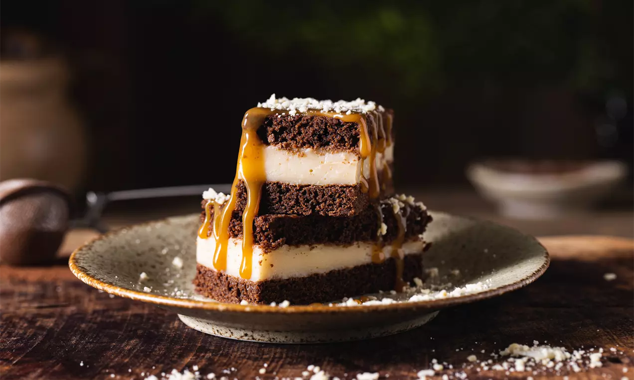 Tmavě hnědé brownie vrstvy s krémově bílým mandlovým pudingem, hustým karamelem a drobnými kousky posypu na rustikálním talíři