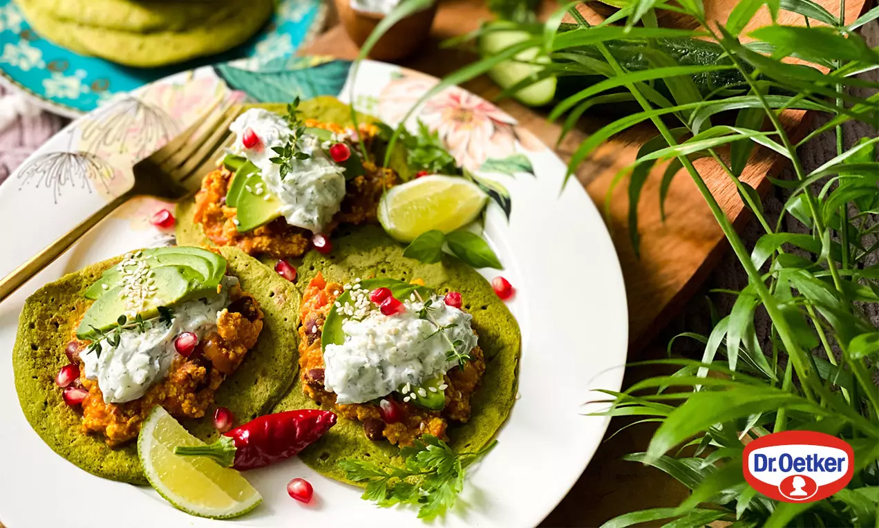 Světle zelené taco placky s hrubší texturou, na nich tofu směs, avokádo, bílý dip, granátové jablko a limetka na zdobeném talíři mezi zelení