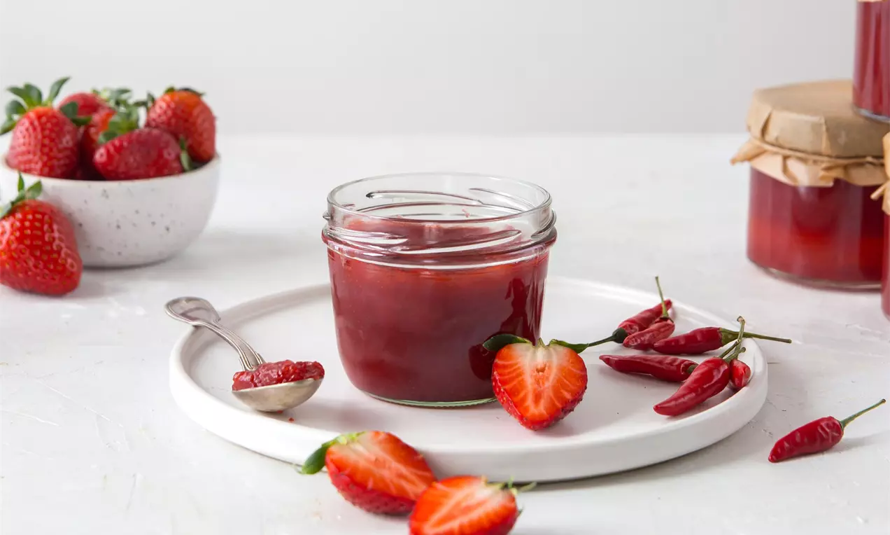 Sklenice tmavě červeného džemu na bílém talíři, obklopená čerstvými jahodami, rozříznutými papričkami chilli a lžící s džemem