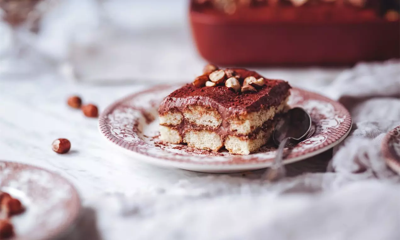Vrstvený dezert prokládaný piškoty, krémovou vrstvou sypaný kakaem a ozdobený ořechy na zdobném dezertním talířku a mramorové desce