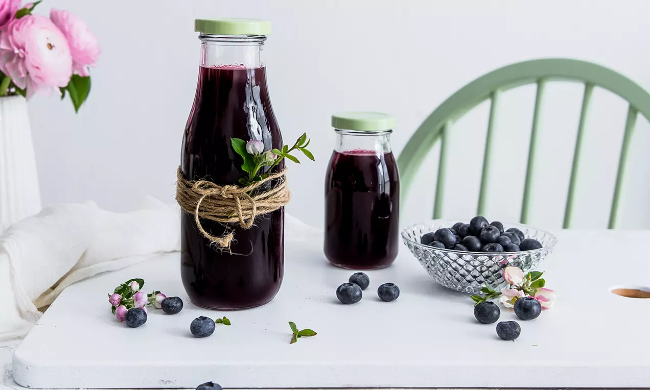 Tmavě fialový borůvkový sirup ve skleněných lahvích s zelenými víčky, ozdobený provázkem a zelení, vedle borůvek v misce na bílém stole