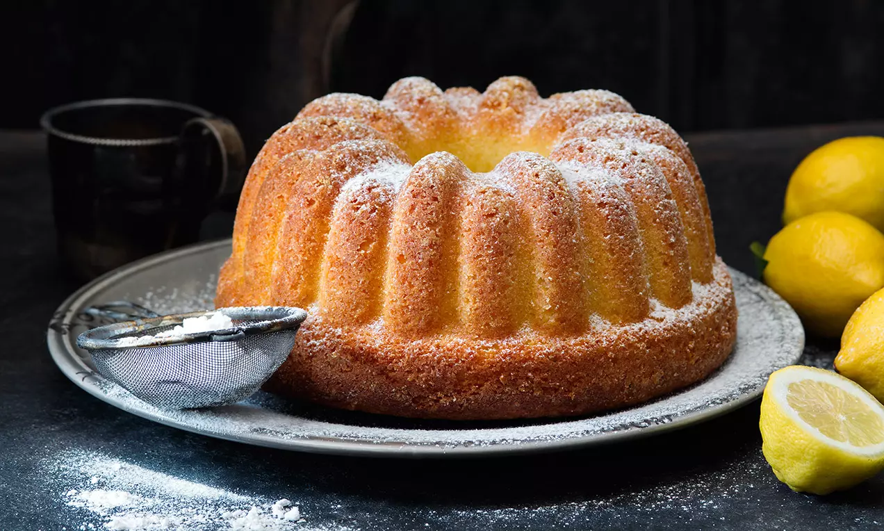 Nadýchaná tvarohová bábovka zlato-hnědé barvy s jemným moučkovým posypem na stříbrném talíři vedle citronů a sítka na tmavém pozadí