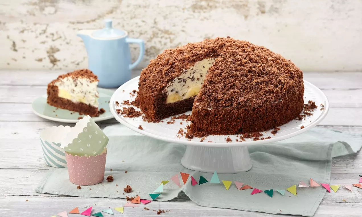 Krtkův dort s tmavě hnědým čokoládovým povrchem, nadýchanou světlou banánovou náplní a drobenkou na bílém talíři s barevnými dekoracemi