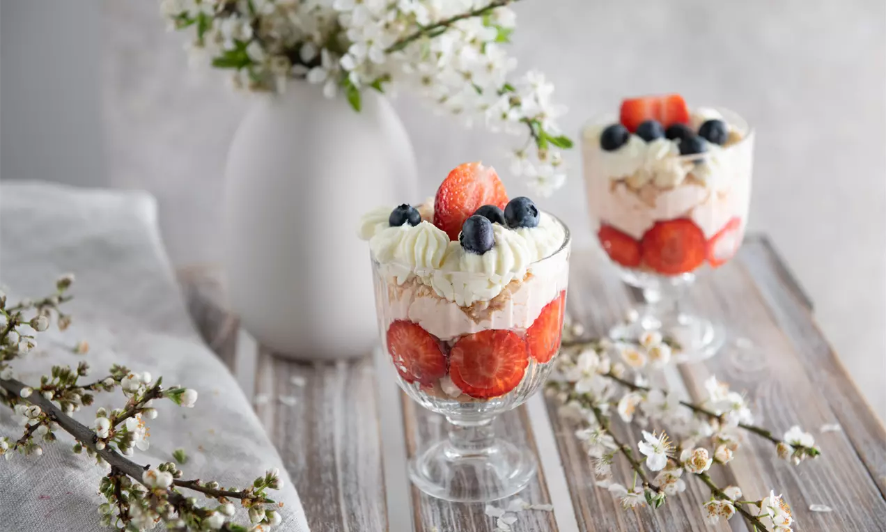 Jahodový pohár v čirém skle s vrstvami krému, mascarpone a šlehačky, ozdobený celými jahodami, borůvkami a větvičkami květů na dřevěném stole