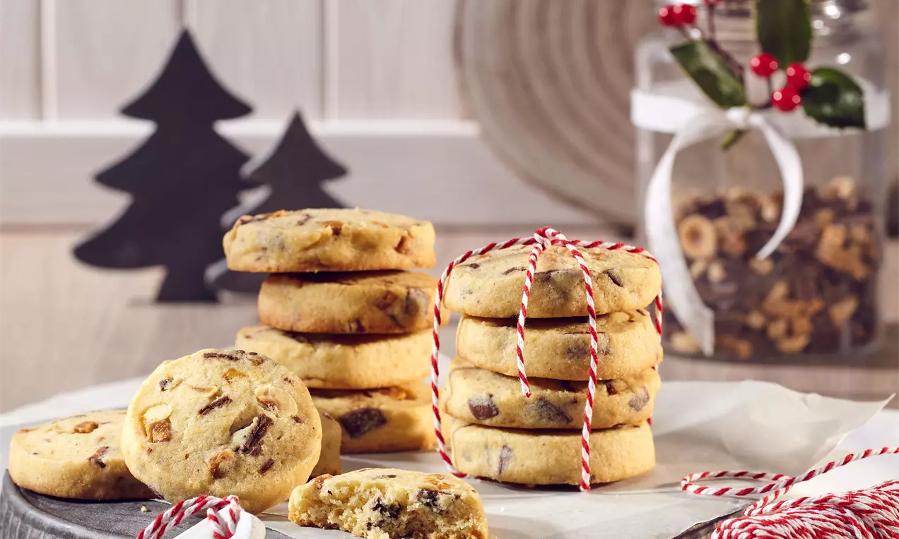 Kulaté cookies světle zlaté barvy s hrubou strukturou, kousky lískových oříšků a karamelu, uspořádané na papíru s ozdobnou šňůrkou