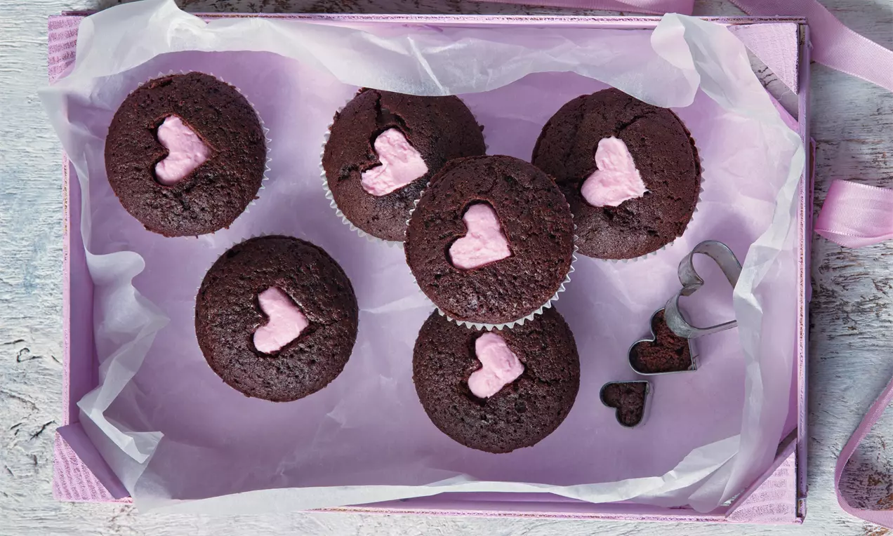 Čokoládové muffiny tmavě hnědé barvy s růžovými srdíčky uprostřed, uspořádané na růžovém podnosu s jemným papírovým zdobením