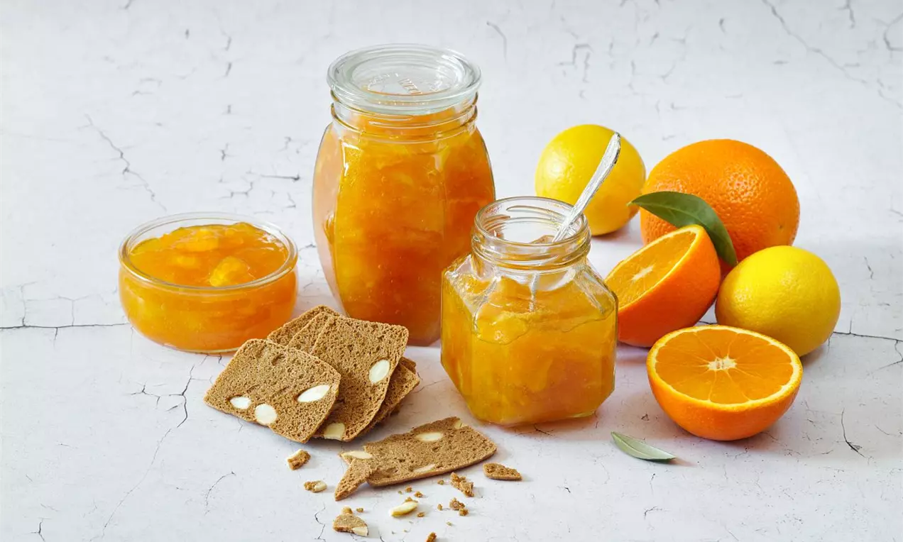 Citrusovo-zázvorová marmeláda sytě oranžové barvy ve sklenicích, s kousky ovoce, obklopená plátky citrusů a křupavými plátky chleba