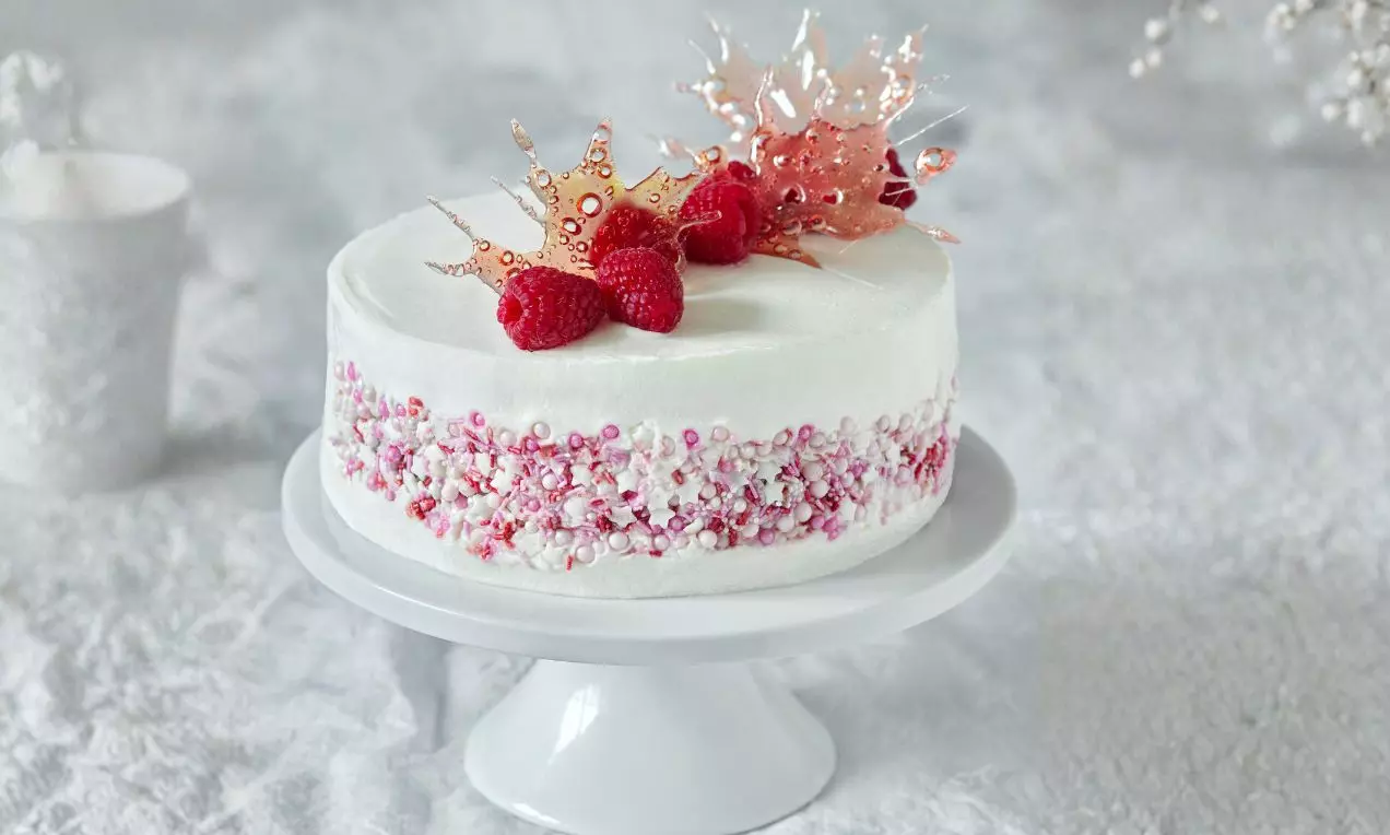 Kulatý dort s nadýchaným bílým krémem, ozdobený malinami, růžovou cukrovou dekorací a perličkami na bílé talíři