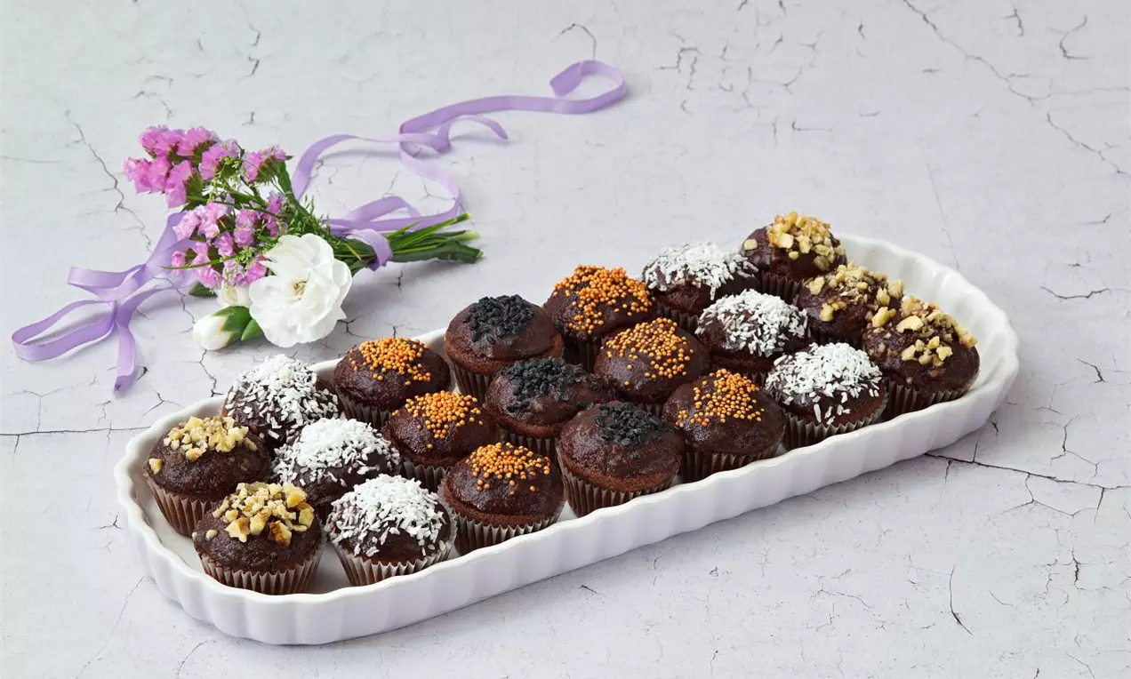 Tmavě hnědé mini muffiny s různými posypy jako kokos, mák, karamel a ořechy na porcelánovém talíři vedle dekorativní květinové vazby