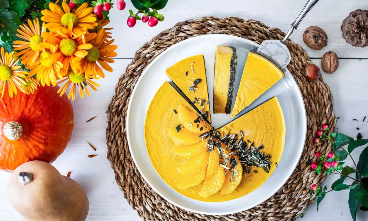 Naříznutý žlutý cheesecake na bílém talířku a proutěném prostírání zdobený dýňovými semínky a filátky z pomeranče s okolní dekorací čerstvými dýněmi a podzimními květy