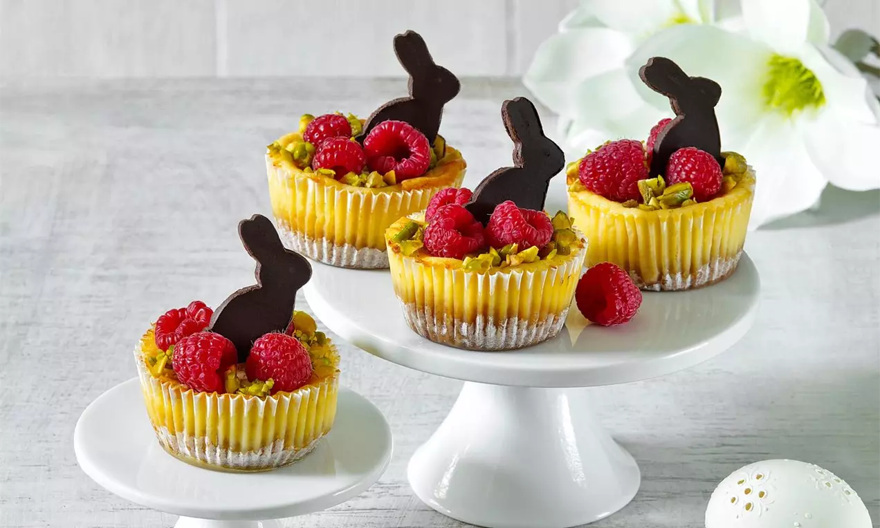 Velikonoční mini cheesecaky s krémovou žlutou náplní v papírových košíčcích, zdobené malinami, pistáciemi a čokoládovými zajíčky na bílých stojanech