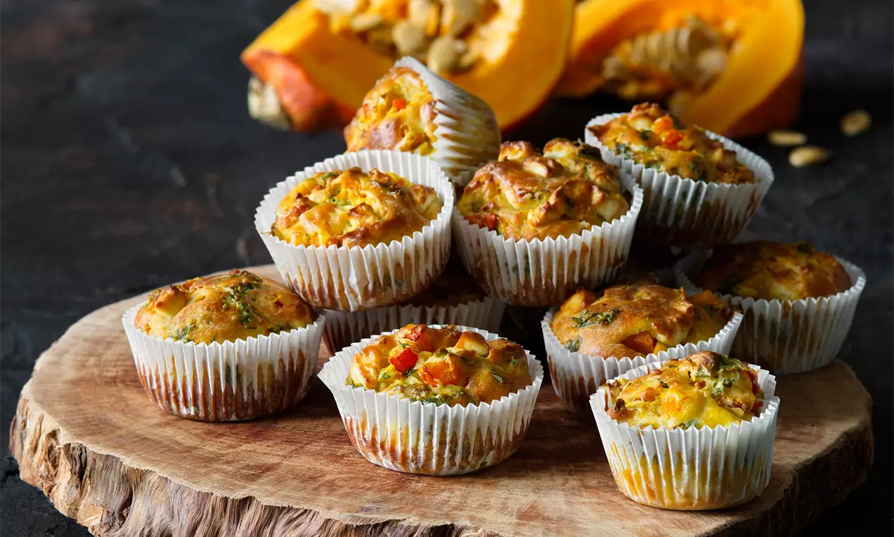 Dýňové slané muffiny zlatohnědé s jemnými oranžovými a zelenými tóny, v papírových košíčcích na dřevěném podnosu s rozkrojenou dýní v pozadí