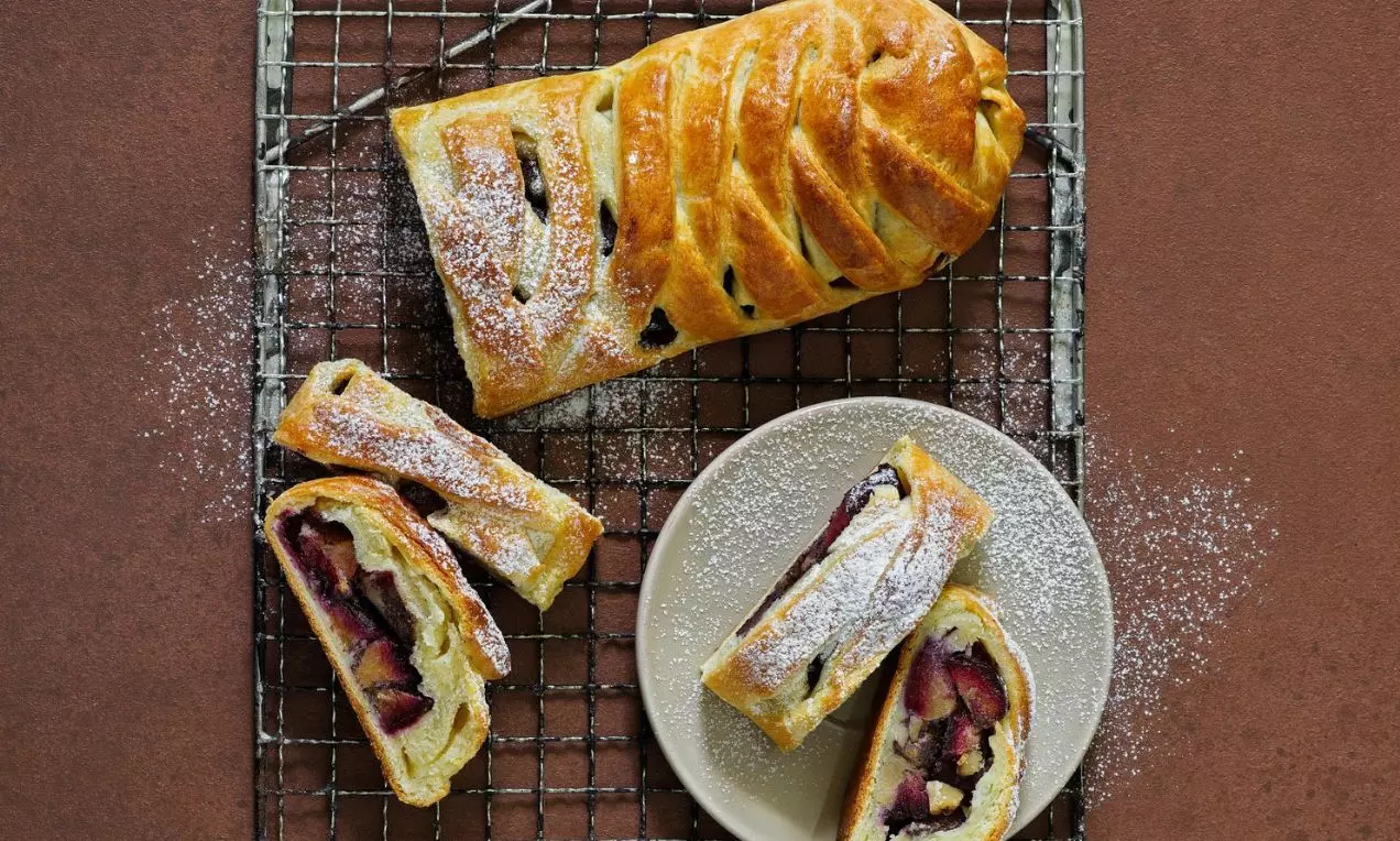 Závin s mákem a švestkami na hnědém stole, pocukrovaný moučkovým cukrem, rozřízlý a servírovaný na bílém talířku