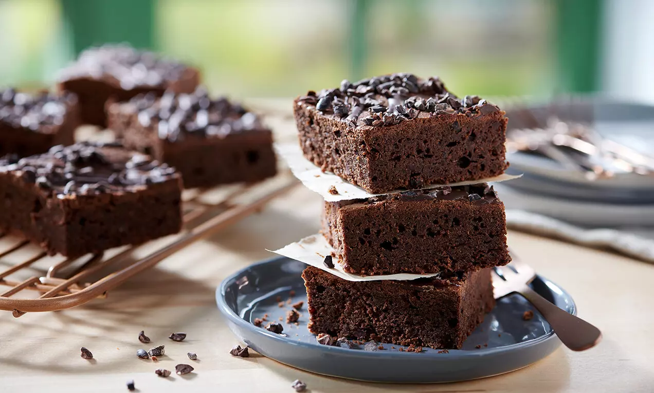 Brownies z červených fazolí sytě hnědé barvy, jemně vlhké uvnitř, posypané drcenými kakaovými boby, vyskládané na modrém talíři s papírem mezi vrstvami