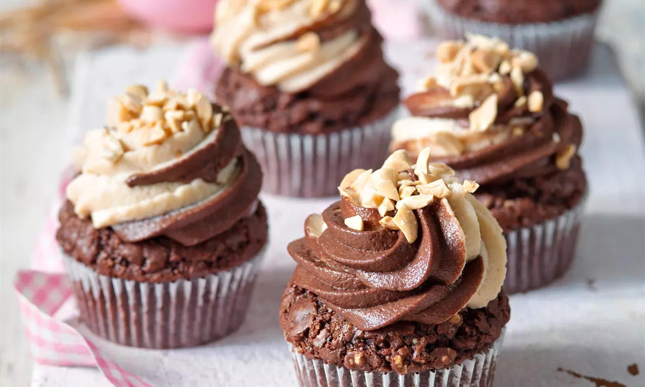 Čokoládové cupcaky s bohatým krémem ve spirálovitých tvarech, posypané nasekanými arašídy, servírované na světlém podkladu s růžovou stuhou