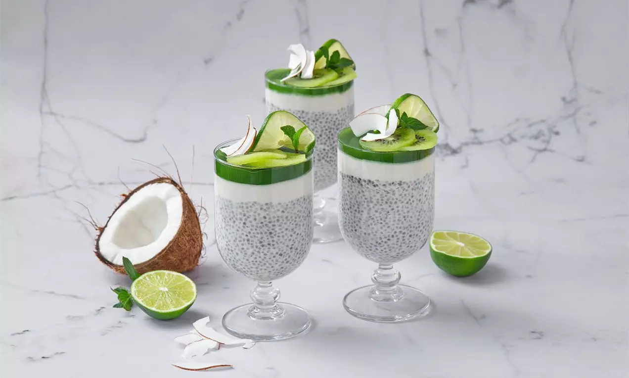Sklenice s chia pudinkem šedé barvy, bílou kokosovou vrstvou a zeleným matcha želé zdobené limetkovým plátkem a lístky máty na mramoru