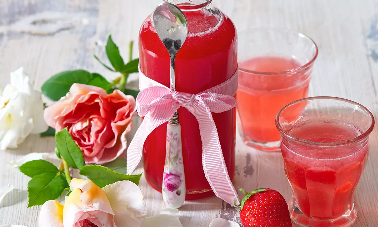 Růžově červený sirup v lahvi s růžovou stuhou a lžičkou, dvě sklenice sirupu, jahoda a růže na světlém dřevěném povrchu