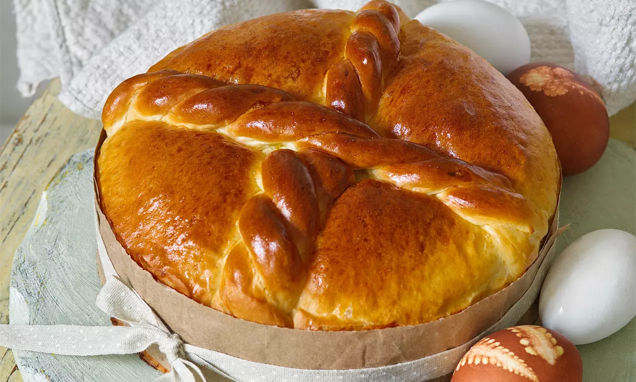Zlatavý kulatý velikonoční chléb s lesklým povrchem a pleteným křížem, položený na přírodním tácku vedle zdobených a bílých vajec