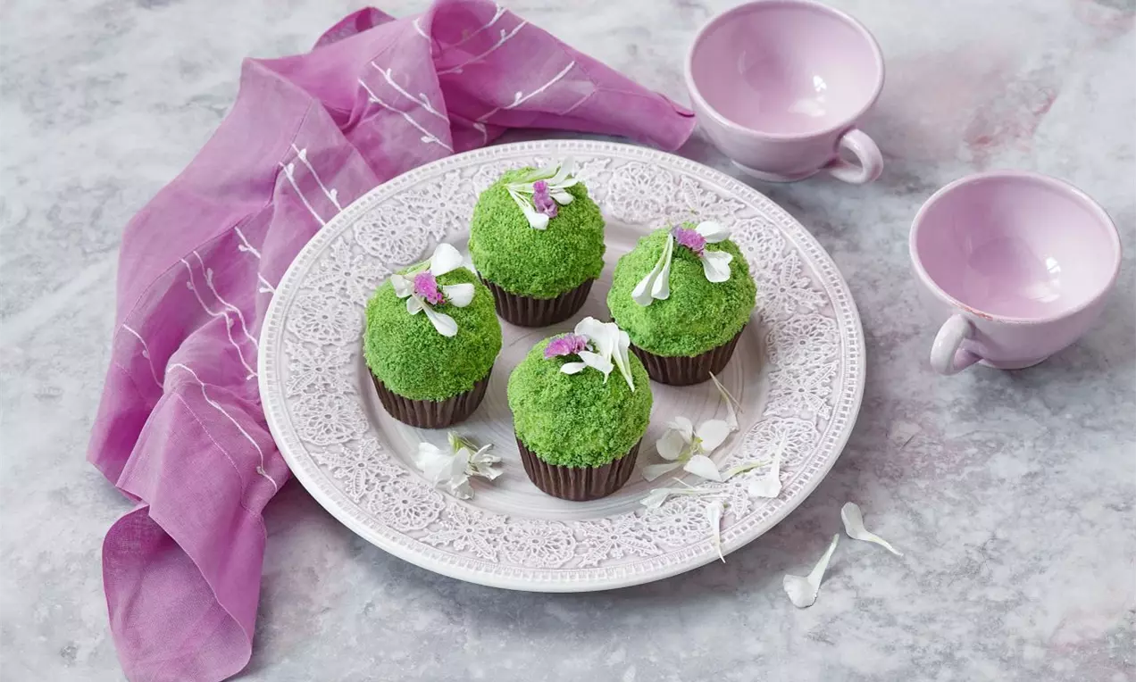 Cupcaky pokryté sytě zelenou drobenkou, ozdobené jemnými kvítky na zdobeném talíři, vedle růžového šátku a porcelánových šálků