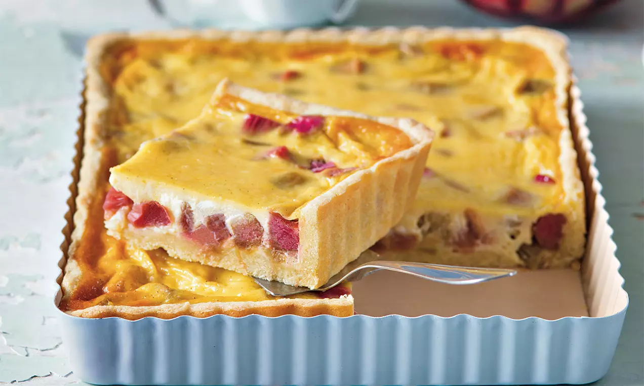 Čtvercový rebarborový koláč se světlou krémovou náplní na zlatavém křupavém korpusu s růžovými kousky rebarbory, servírovaný na stříbrné lžíci