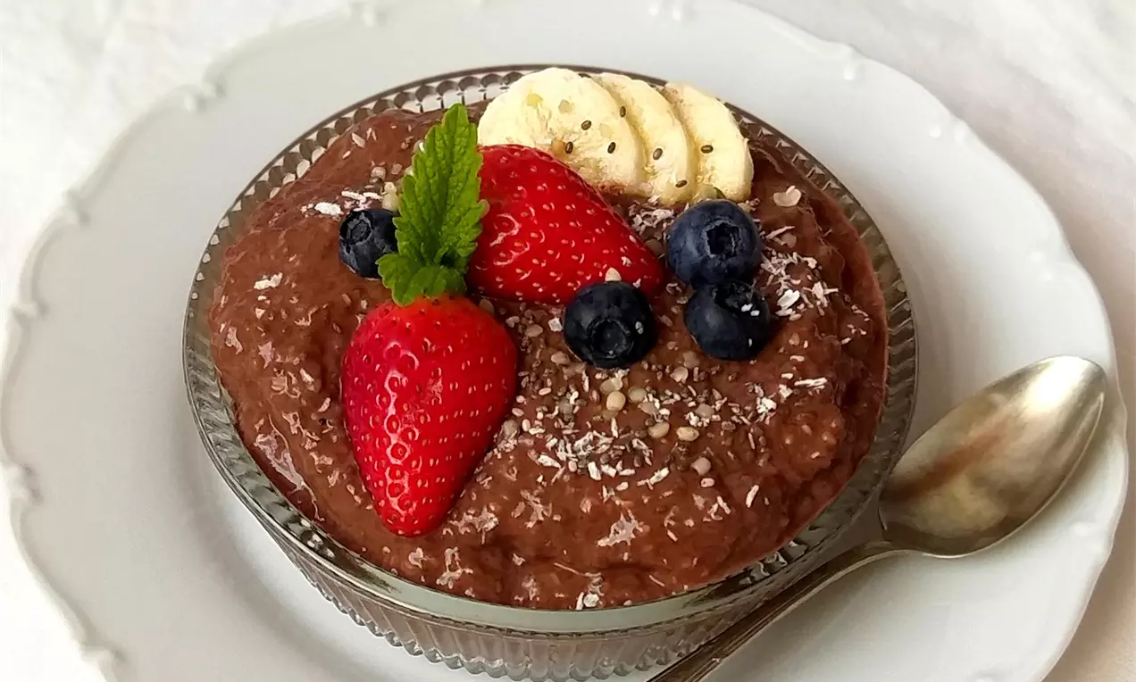 Hustý čokoládový pudink tmavé barvy v skleněné misce, zdobený jahodami, borůvkami, banánem, mátovým lístkem a strouhaným kokosem na bílém talíři
