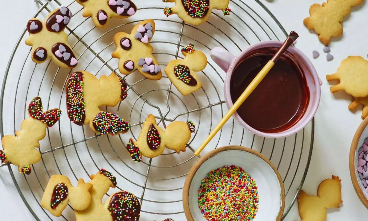 Velikonoční sušenky ve tvaru zvířátek a vajíček, zlatavé pečivo s barevnou čokoládovou polevou a posypem na kovové mřížce vedle misek s dekoracemi
