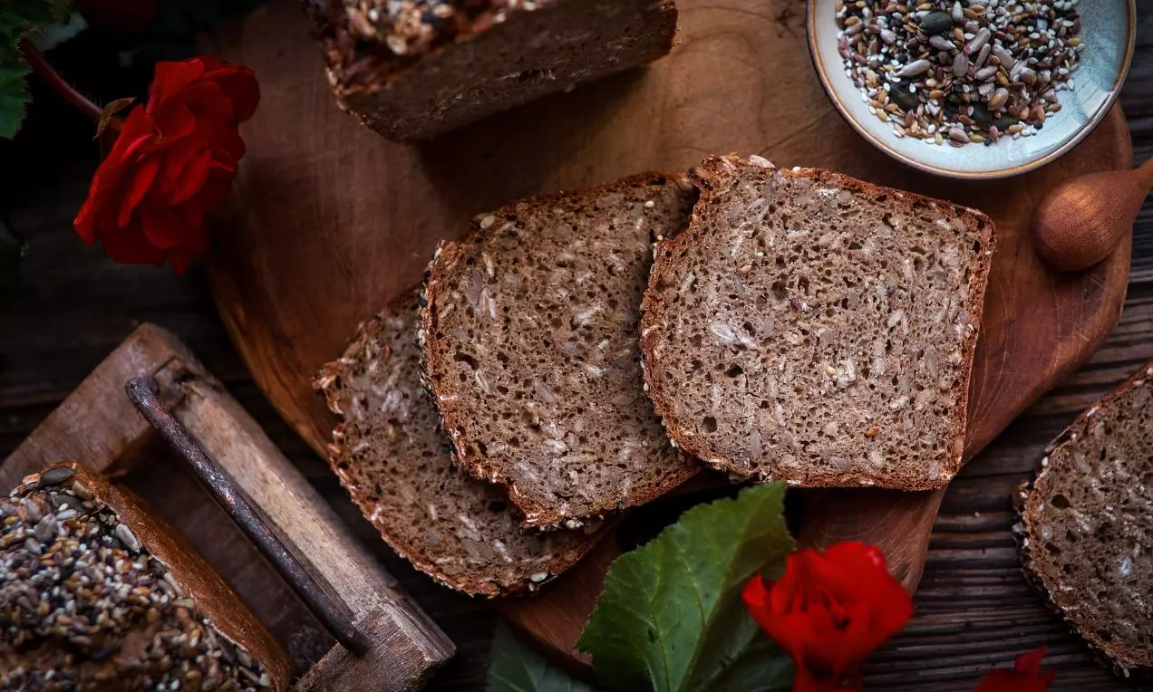 Tmavý žitný kváskový chléb na dřevěném prkénku, plátky s hrubou strukturou, vedle mísa semínek a červené květy jako dekorace
