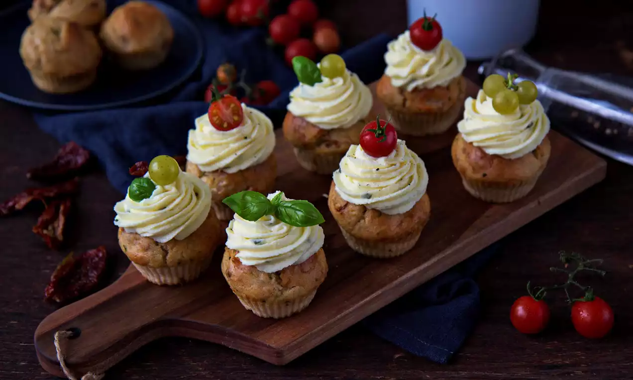 Slané muffiny zlatohnědé barvy s krémovým sýrovým toppingem, ozdobené čerstvými cherry rajčátky, bobulemi hroznů a bazalkou na dřevěné desce