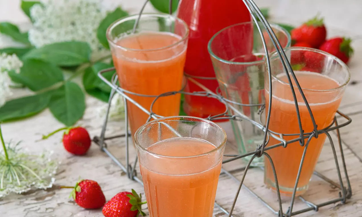 Světle růžový sirup ve sklenicích v kovovém nosiči, doplněný lahví a dekorací z čerstvých jahod a květů bezinky na světlém dřevěném povrchu