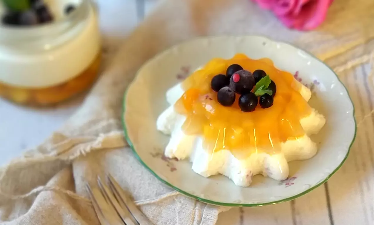 Panna cotta ve tvaru vějířovitého květu s jemnou bílou texturou a hladkým mangovým želé, zdobená lesklými bobulemi na květovaném talíři